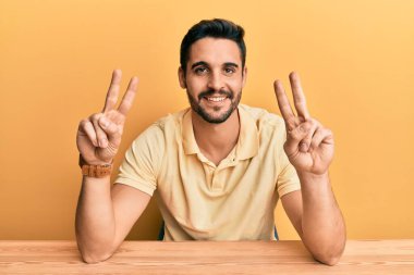 Gündelik giysiler giymiş genç İspanyol adam masaya oturmuş gülümsüyor ve parmaklarını zafer işareti yaparken kameraya bakıyor. İki numara.. 