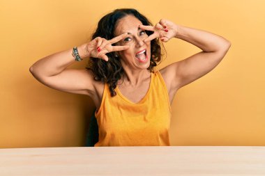 Masaya oturmuş barış sembolü yapan orta yaşlı, güzel bir kadın. Yüzü parmaklarıyla gülümsüyor ve zafere gülümsüyor. 