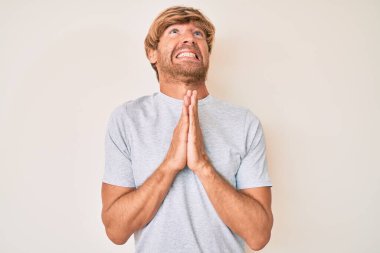 Sıradan kıyafetler giyen genç sarışın adam elleri bağlı yalvarıyor ve dua ediyor. Yüzünde umut ifadesi var. Çok duygusal ve endişeli. Yalvarıyor. 