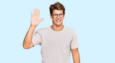 Yakışıklı beyaz adam, günlük kıyafetler ve gözlük takıyor. Beş numarayı gösteriyor ve işaret ediyor. Kendinden emin ve mutlu gülümsüyor.. 