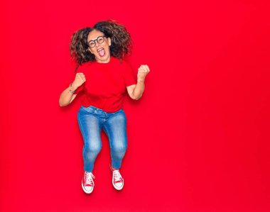 Orta yaşlı, güzel İspanyol bir kadın günlük kıyafetler ve mutlu bir gülümseme takınıyor. İzole edilmiş kırmızı arka planda yüzünde gülümsemeyle zıplıyor