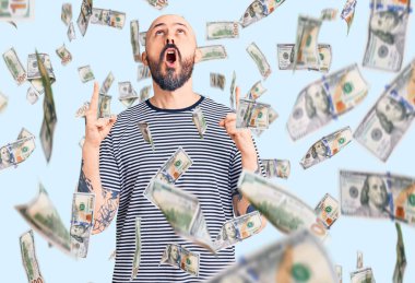 Gündelik giysiler giyen genç yakışıklı adam şaşırmış ve şaşırmış bir şekilde yukarı bakıyor ve parmaklarıyla işaret ediyor ve kollarını kaldırıyor..