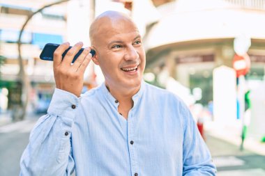 Orta yaşlı kel adam gülümsüyor. Şehirdeki akıllı telefonu kullanarak mutlu bir sesli mesaj dinliyor..