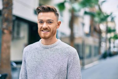 Genç beyaz adam gülümseyerek şehirde mutlu mesut duruyor..