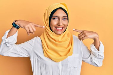 Genç esmer Arap kadın geleneksel İslami başörtüsü takıyor neşeli gülümsemeler sergiliyor ve parmaklar, dişler ve ağızla işaret ediyor. diş sağlığı kavramı. 