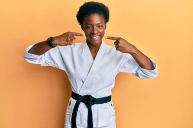 Karate kimonosu ve siyah kuşak giyen, gülümseyen, parmaklarını ve ağzını gösteren Afro-Amerikalı genç bir kız. diş sağlığı kavramı. 