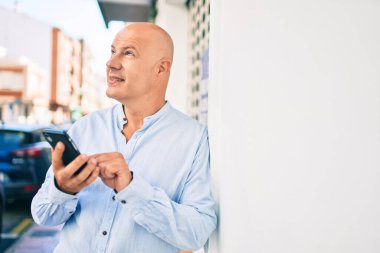 Orta yaşlı kel adam şehirde akıllı telefon kullanarak mutlu bir şekilde gülümsüyor..