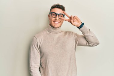 Balıkçı yaka kazak giyen İspanyol genç adam barış sembolü yapıyor. Yüzünde parmaklar, gülümseyerek zaferi gösteriyor. 