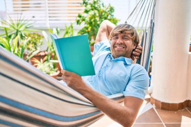 Orta yaşlı yakışıklı bir adam evinin terasında bir hamakta uzanıp kitap okuyor.