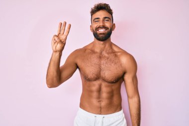 Genç İspanyol adam üstsüz duruyor ve kendini beğenmiş ve mutlu bir şekilde gülümserken üç numarayı işaret ediyor.. 