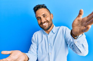 Sakallı, sıradan kıyafetler giyen yakışıklı kameraya sarılıp gülümseyen bir adam. Mutluluğu kucaklayan neşeli bir ifade.. 