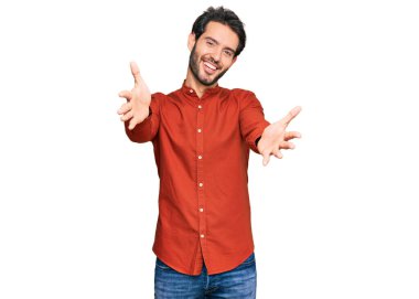 Gündelik giysiler giymiş genç İspanyol adam kameraya bakıp sarılmak için kollarını açmış gülümsüyor. Mutluluğu kucaklayan neşeli bir ifade.. 