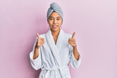 Duş havlusu şapkası ve bornoz giyen genç Çinli kadın el hareketleriyle olumlu bir jest yapıyor, gülümsüyor ve mutlu. Neşeli ifade ve kazanan jest. 