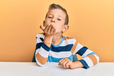 Sevimli beyaz çocuk, gündelik kıyafetlerini giymiş masaya oturmuş kameraya bakıyor ve havada el ele öpüşüp sevimli ve seksi oluyor. aşk ifadesi. 