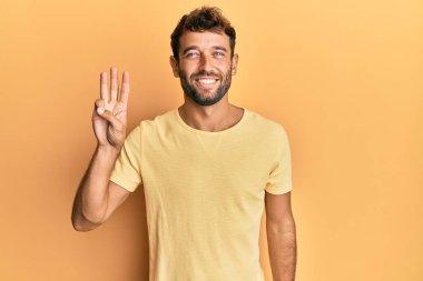 Sarı arka planda sarı tişört giyen sakallı yakışıklı adam üç numaralı parmaklarıyla gülümseyerek kendinden emin ve mutlu bir şekilde onu işaret ediyor.. 