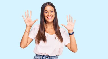 Genç İspanyol kadın, günlük beyaz tişört giyer ve parmaklarını on numarayla gösterip gösterirken kendinden emin ve mutlu bir şekilde gülümser.. 