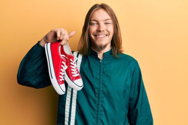 Uzun saçlı, kırmızı ayakkabılı yakışıklı beyaz bir adam. Pozitif ve mutlu görünüyor. Kendine güvenen bir gülümsemeyle dişlerini gösteriyor. 