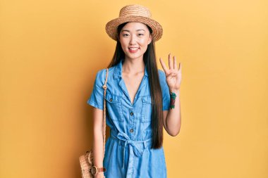 Genç Çinli kadın yaz şapkası takıyor ve parmaklarıyla dört numarayı gösterirken kendinden emin ve mutlu bir şekilde gülümsüyor.. 