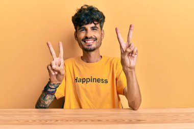 Üzerinde mutluluk mesajı olan tişört giyen genç İspanyol adam masaya oturmuş gülümsüyor ve parmaklarını zafer işareti yaparken kameraya bakıyor. İki numara.. 