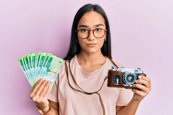 Young asian woman holding vintage camera and 200 russian ruble banknotes relaxed with serious expression on face. simple and natural looking at the camera.