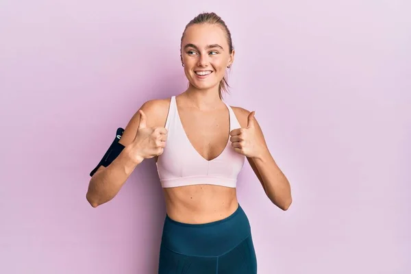 Beautiful blonde woman wearing sportswear and arm band success sign doing positive gesture with hand, thumbs up smiling and happy. cheerful expression and winner gesture.