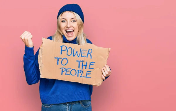 Young caucasian woman holding power to the people banner screaming proud, celebrating victory and success very excited with raised arms