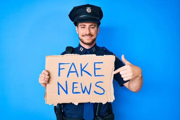 Young caucasian man wearing police uniform holding fake news banner smiling happy pointing with hand and finger