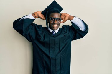 Yakışıklı siyah adam mezuniyet şapkası ve tören cübbesi takıyor rahatlıyor ve esniyor, kolları ve elleri başının arkasında mutlu bir şekilde gülümsüyor. 