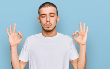 Latin asıllı beyaz tişört giyen genç adam rahatla ve gözleri kapalı gülümse parmaklarıyla meditasyon hareketi yapıyor. yoga konsepti. 