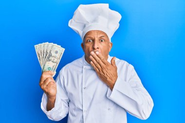 Orta yaşlı, gri saçlı, profesyonel aşçı üniforması giyen, elinde Amerikan doları banknotları tutan ağzı ağızla kaplayan, şok olmuş ve hata yapmaktan korkan bir adam. şaşırmış ifade 