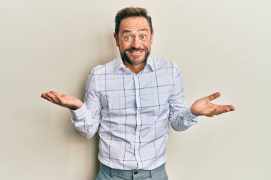 Orta yaşlı bir adam iş kıyafetleri giyiyor ve elleri ve kolları havada, kafası karışmış bir ifade takınıyor. şüphe kavramı. 