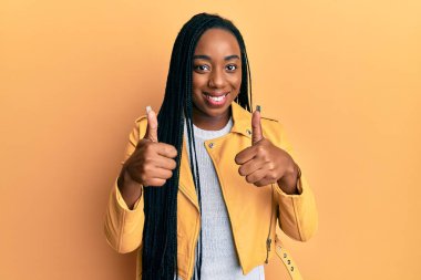Genç Afro-Amerikalı kadın, günlük ceket giyen, başarı işareti yapan, el hareketi yapan, gülümseyen ve mutlu. Neşeli ifade ve kazanan jest. 