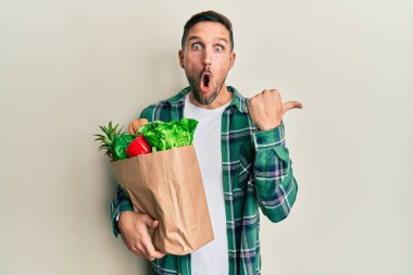 Sakallı yakışıklı bir adam, elinde poşetle bakkaliyeyi gösterirken şaşırmış, ağzı açık, ağzı açık, hayrete düşmüş.. 