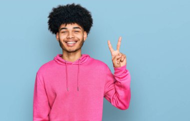 Afro saçlı, sıradan pembe kazak giyen genç Afro-Amerikalı adam iki numarayı gösterirken kendine güvenen ve mutlu bir şekilde gülümsüyor.. 