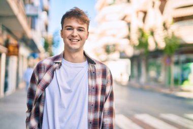 Genç beyaz adam gülümseyerek şehirde mutlu mesut duruyor..