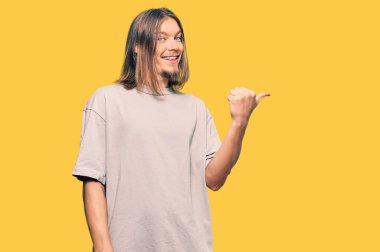 Uzun saçlı yakışıklı, sıradan kıyafetler giyen, gülen yüzlü gülümseyen ve baş parmağıyla yüzünü gösteren beyaz bir adam.. 