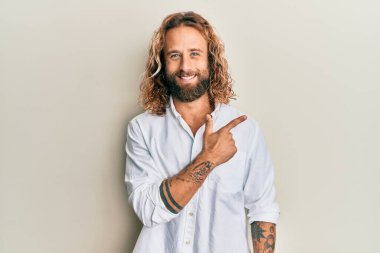 Sakallı, uzun saçlı, sıradan beyaz tişörtlü, neşeli, eli ve parmağı yana doğru uzanan yakışıklı bir adam. 