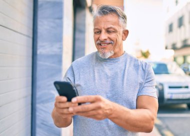 Orta yaşlı, ispanyol, kır saçlı, mutlu gülümseyen adam şehirde akıllı telefon kullanıyor..