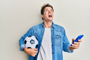 Yakışıklı beyaz adam elinde futbol topu ile akıllı telefona bakıyor kızgın ve kızgın bağırıyor sinirli ve öfkeli, öfkeyle bağırıyor.. 