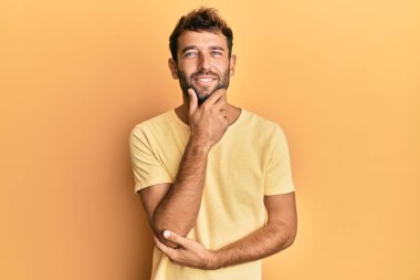 Sarı arka planda sarı tişört giyen sakallı yakışıklı adam kameraya kollarını kavuşturup gülümseyen ve çene üzerinde el kaldıran adam. Olumlu düşünmek. 