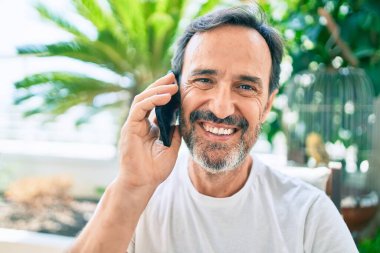 Orta yaşlı, sakallı, gülümseyen, telefonda mutlu konuşan bir adam.