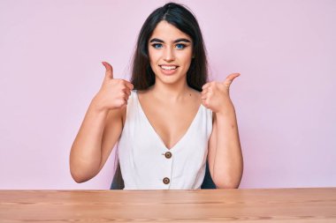 Esmer genç kız gündelik giysiler içinde masaya oturmuş mutlu ve pozitif gülümsüyor, başparmağıyla mükemmel ve onay işareti yapıyor. 