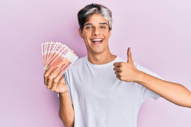 Genç İspanyol adam elinde 100 Norveçli kronu tutuyor mutlu ve olumlu gülümsüyor, başparmağıyla mükemmel ve onay işareti yapıyor. 