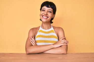 Masanın üzerinde oturan güzel esmer kadın. Sarı arka planda mutlu bir yüz. Kollarını kavuşturup kameraya bakıyor. pozitif kişi. 