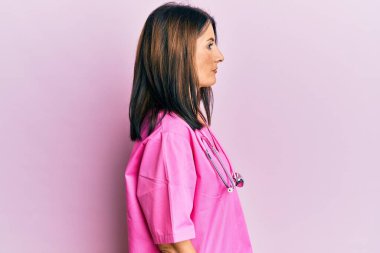 Orta yaşlı esmer kadın, doktor üniforması ve steteskop giyiyor. Yan tarafa bakıyor, rahat profil pozu veriyor, kendine güvenen bir gülümsemesi var.. 