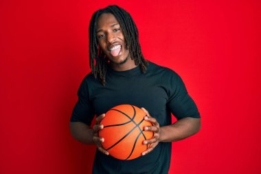 Saçları örgülü, basketbol topu tutan, dilini dışarı çıkaran Afrikalı Amerikalı bir adam. Komik bir ifadeyle mutlu.. 
