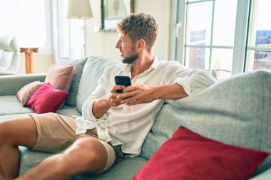 Yakışıklı beyaz adam mutlu gülümsüyor. Evdeki kanepede otururken akıllı telefon kullanıyor.