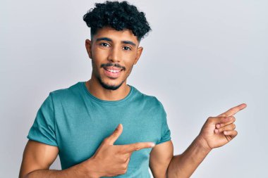 Genç Arap yakışıklı, günlük kıyafetler giymiş, gülümseyen ve kameraya bakan iki eli ve parmakları yandan gösteren adam.. 