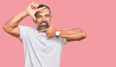 Orta yaşlı, beyaz tişört giyen yakışıklı bir adam gülümseyerek elleri ve parmakları gülen yüzlü bir çerçeve yapıyor. yaratıcılık ve fotoğrafçılık kavramı. 