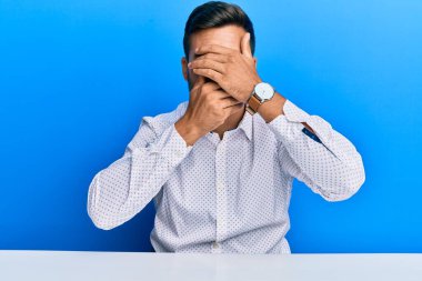 İş kıyafetleri giymiş yakışıklı İspanyol bir adam masanın üzerinde oturuyor gözlerini ve ağzını elleriyle kapatıyor, şaşırmış ve şok olmuş. duygu gizleme 